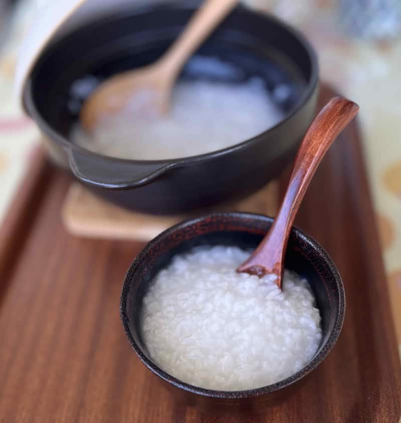 土鍋が背景のお椀に真っ白なお粥がよそってあり木製のスプーンが入っている
